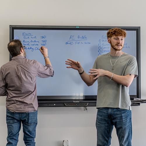 Student member speaking in front of AI Club members while Professor Robert Kissner writes on white board.
