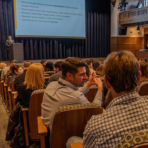 Audience members talk in pairs while a presentation slide on innovation is displayed behind the speaker.