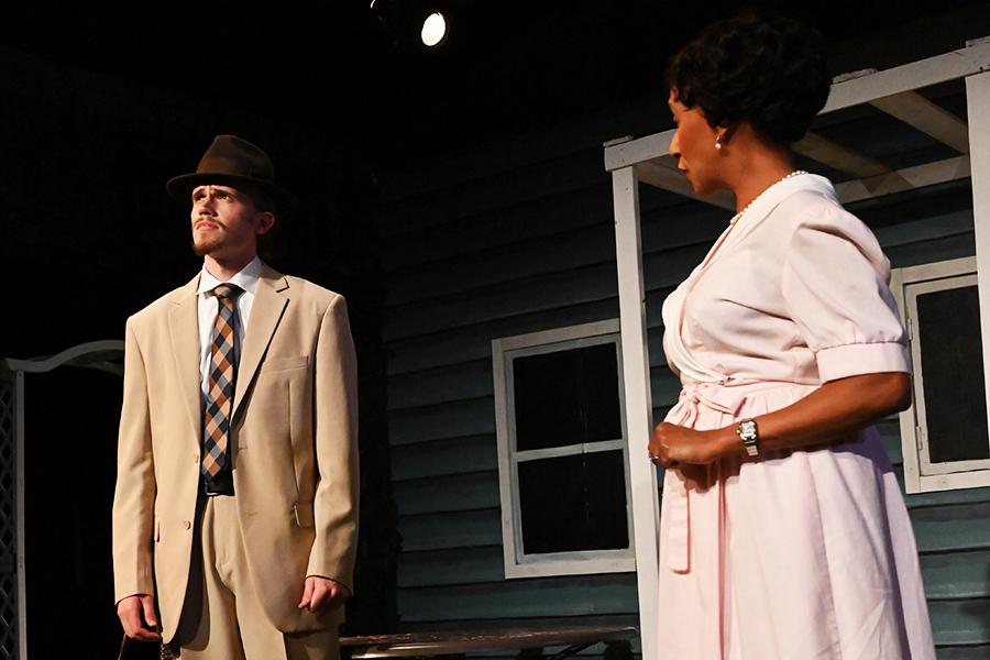 A man in a suit and hat looks away while a woman speaks to him.
