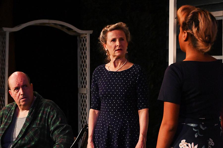 Two women speak to each other while a man in a bathrobe sits at a table and listens.