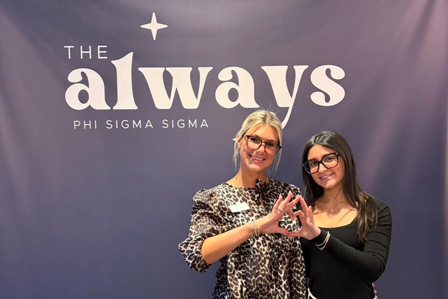 Two members of Phi Sig with a backdrop that says The Always Phi Sigma Sigma.
