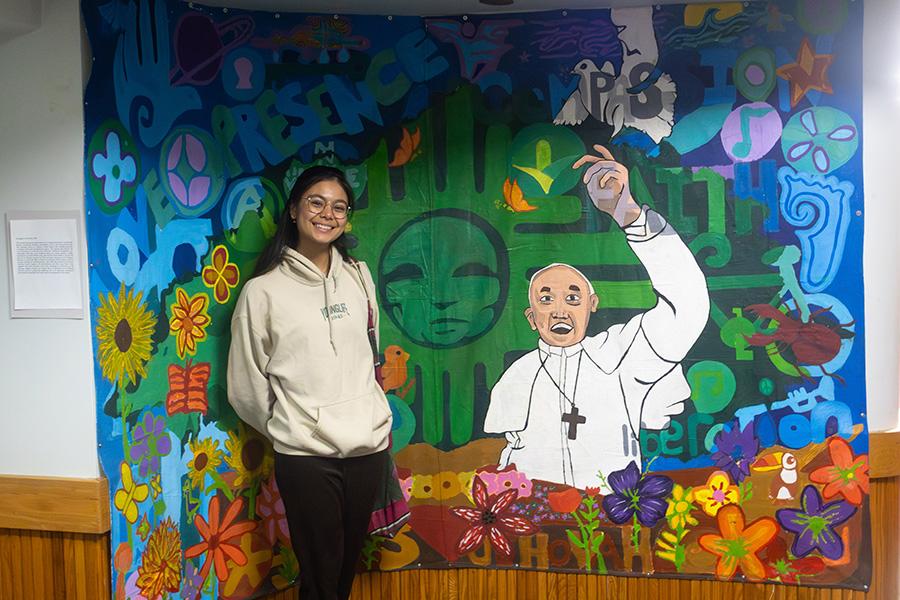Jidapha Vastola ’27 standing and smiling next to the mural titled “Ecological Conversion.”