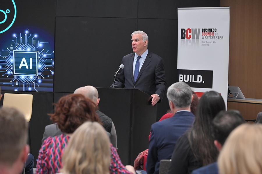John Ravitz, COO of the Business Council of Westchester, stands at a black lectern speaking to an audience at the AI Alliance 360° Impact Summit.