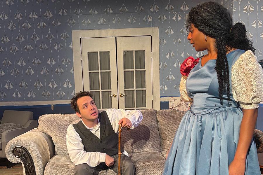 A male actor sitting on the couch, leaning on his cane, looking up to a female actor standing, looking down to the male actor, in the play "Creditors."