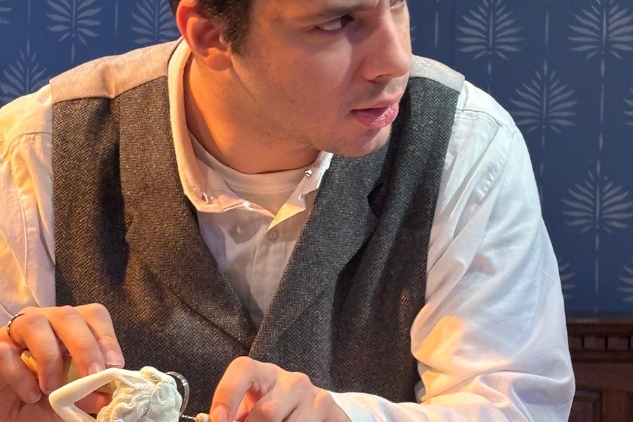 A male actor looking off to his left side, while his hands hold steady as he's sculpting a white marble statue of a woman.