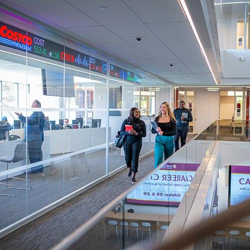 Three students are in the LaPenta School of Business, walking down the hallway, passing by the LaPenta-Lynch Trading Floor on their left.