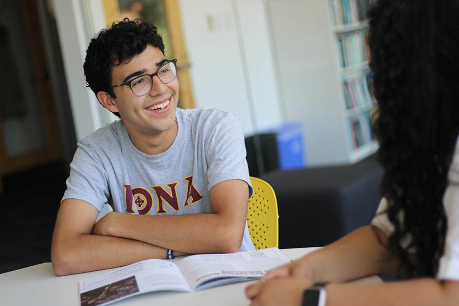 Student sitting and smiling in a classroom while conversing with a professor.