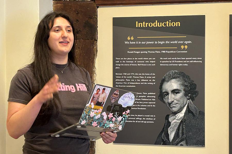 A student talking and holding a laptop, in front of a poster of Thomas Paine.
