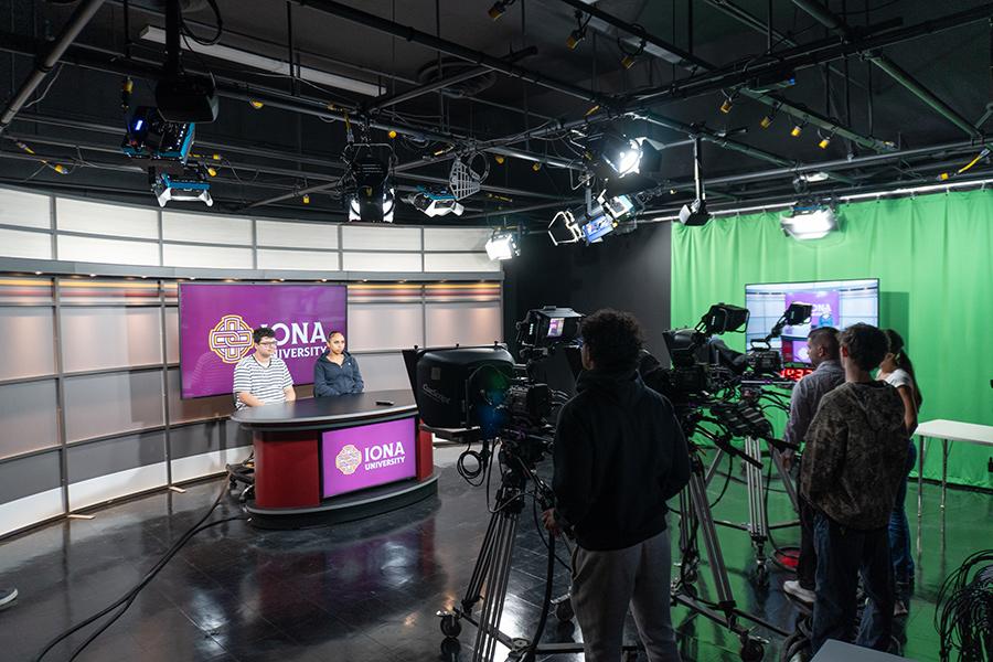 Two students sitting at the broadcast table, while three other students are working the cameras with the faculty member teaching everyone.