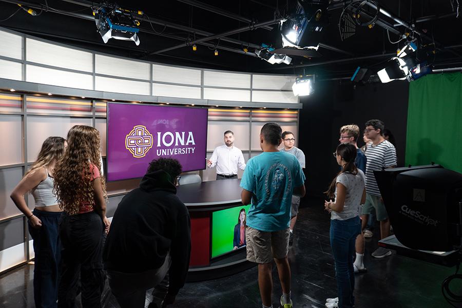 Students gathered around the broadcast table, listening to faculty member speak.