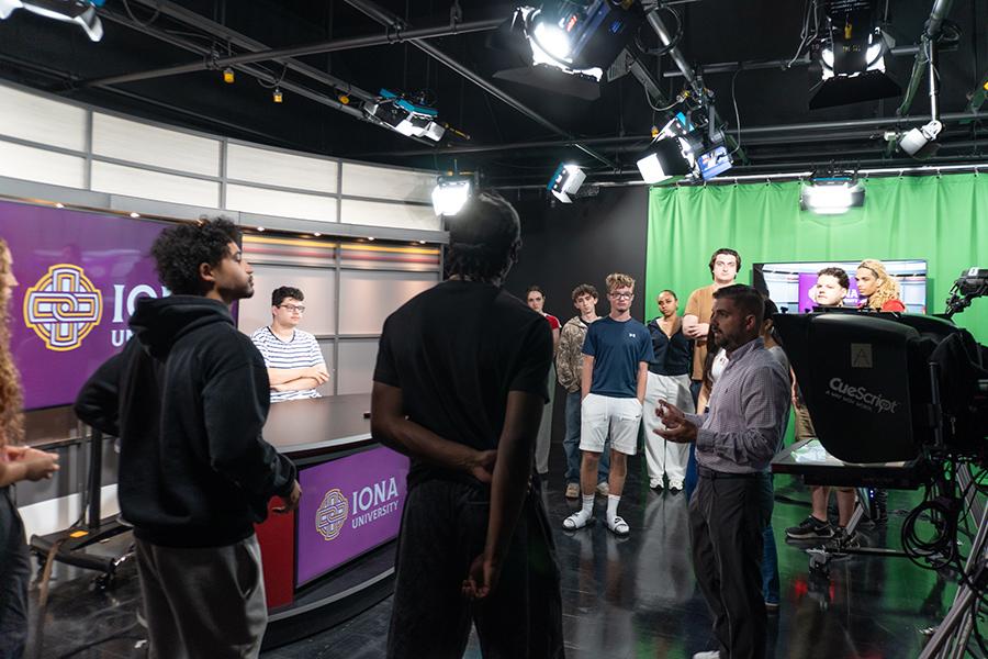 Several students standing in the broadcast room, learning about the Iona University Television set up from a faculty member.