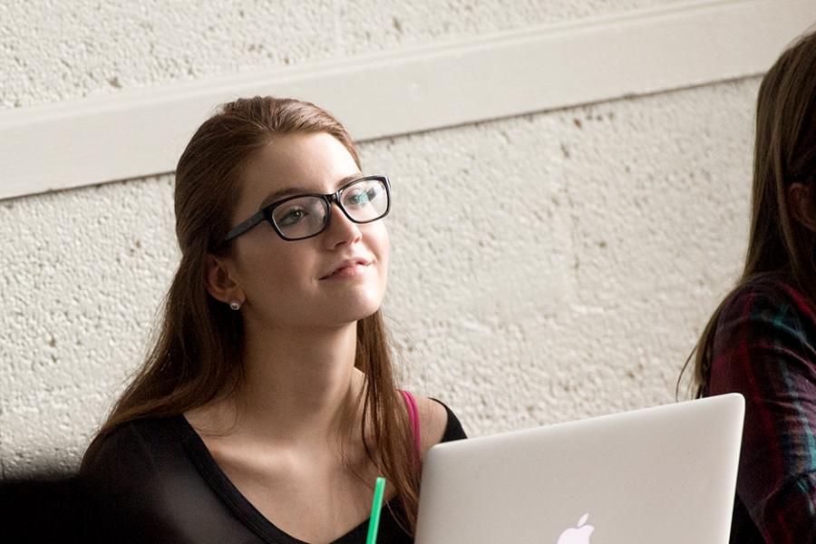 A student with glasses has her laptop in class and smiles and looks toward the professor.