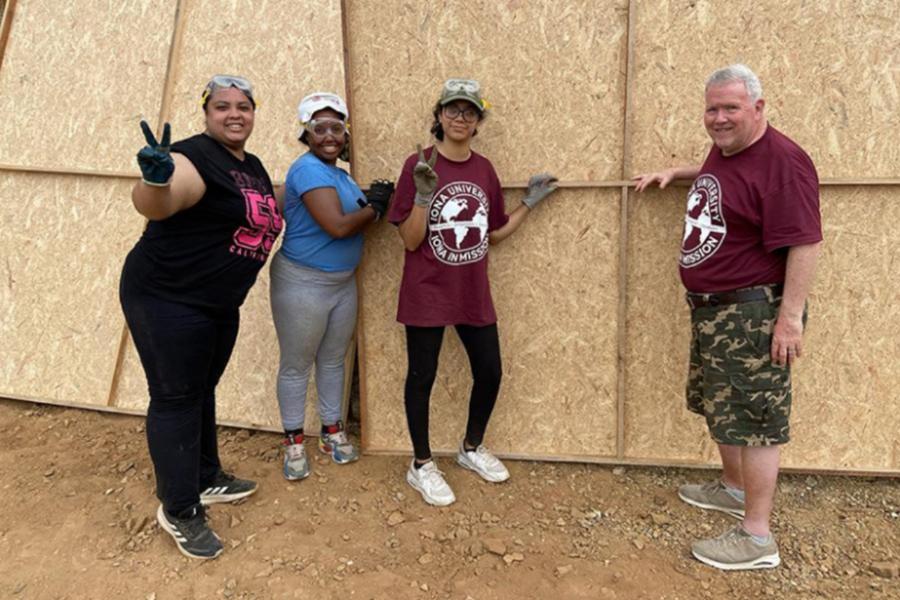 Three Iona students with Father Gerard, taking a group photo in front of wood panels.