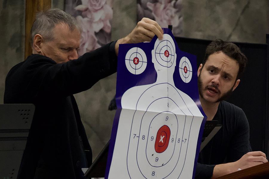 Two male actors, during the performance of "Motherland," one male actor is holding a paper target while the other male actor looks on.
