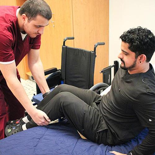 Two OT students practice assisting a client get into bed from a wheelchair.
