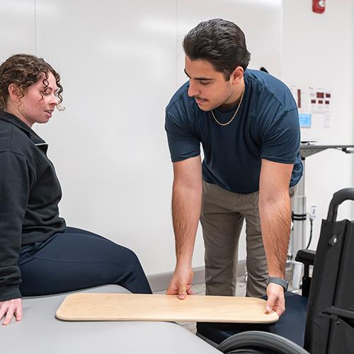 Two OT students practice helping a patient from the bed to a wheelchair.