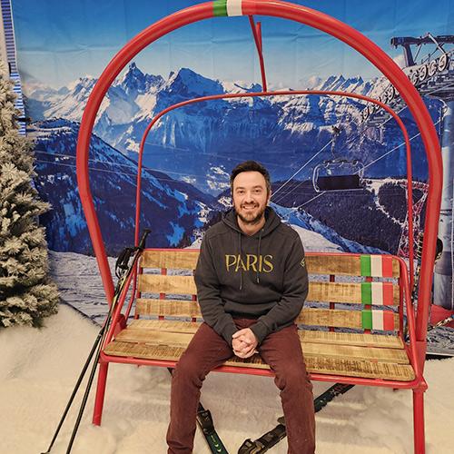 Allen Pines ’21MA smiles while sitting on a red ski lift bench in front of a winter mountain backdrop.