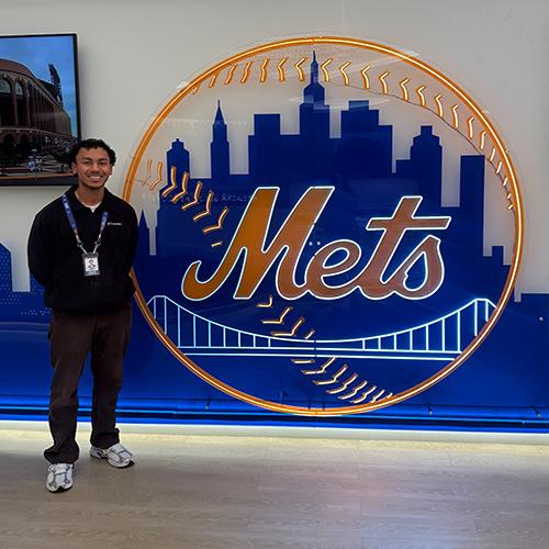 Diego Aguilar standing and posing in front of Mets logo sign during Mets internship.
