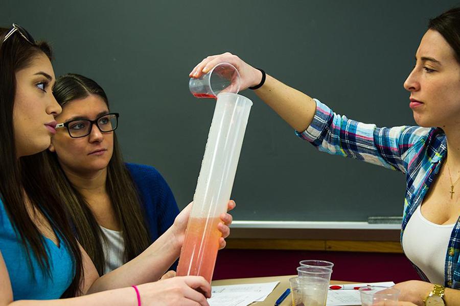 Three students sit at a table in chemistry class. One pours liquid into a cylinder and another holds the cylinder.