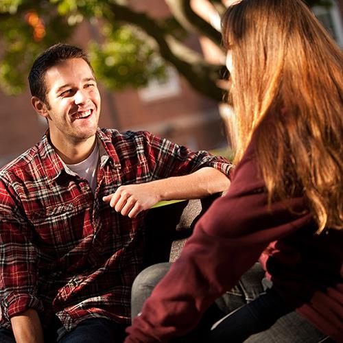 A student in a flannel shirt sits on a bench and talks with another student.