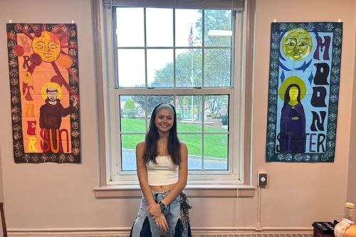 Jidapha Vastola ’27 standing and smiling in the middle of the two individual banners which is entitled “Brother Sun, Sister Moon."