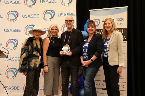 Dr. Christoph Winkler stands in front of a USASBE backdrop while holding his 2026 Entrepreneurship Educator of the Year award.