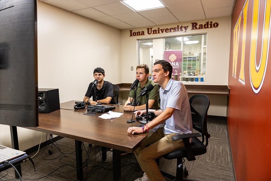 Three male students, in the Iona University Radio Station Room, watching the screen and doing a live radio broadcast.