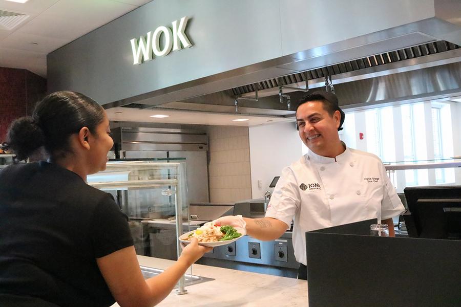 A chef hands a plate of food to a student from the Wok station.